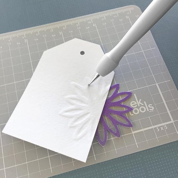 White paper cutout with a purple flower design on a cutting mat with a tool.