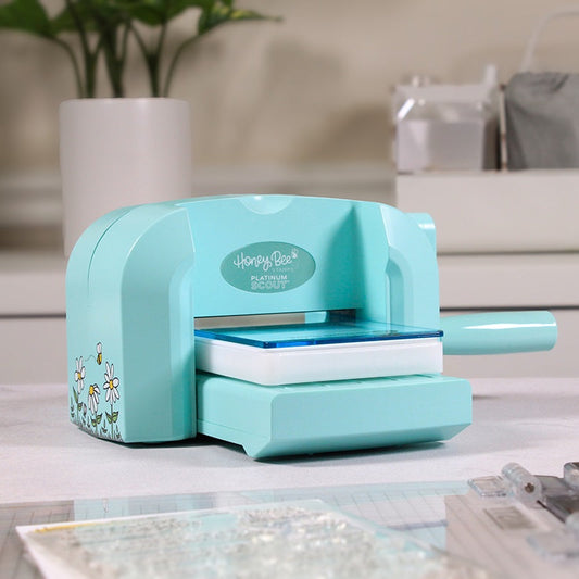 A turquoise paper cutter machine with floral accents sits on a light countertop in a kitchen or craft workspace, with blurred cabinets, a plant, and kitchen items in the background.