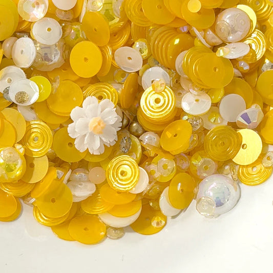 A pile of yellow and white craft sequins, beads, and small decorative flower pieces on a plain white background.