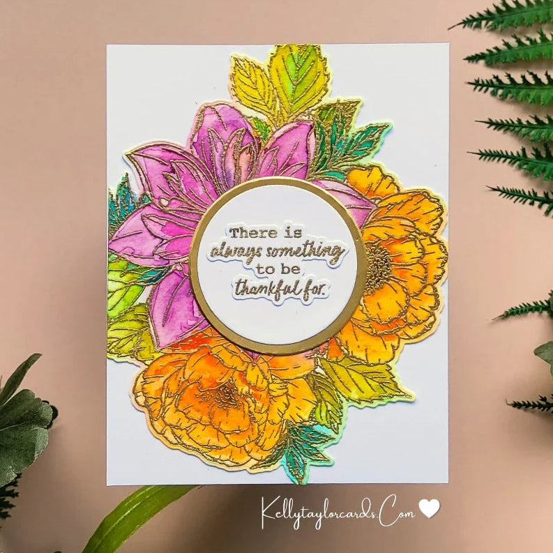 A handmade greeting card featuring colorful floral illustrations in pink, orange, yellow, and green, with a central gold-bordered circle containing the hand-lettered quote 'there is always something to be thankful for.' The card is displayed on a beige background with decorative green leaves and white flowers around it.