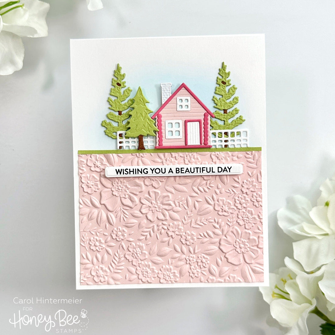 Handmade greeting card featuring a pink cabin, green trees, and a floral embossed background.