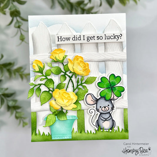 Handmade card featuring a grey mouse with a four-leaf clover and yellow roses by a white fence.