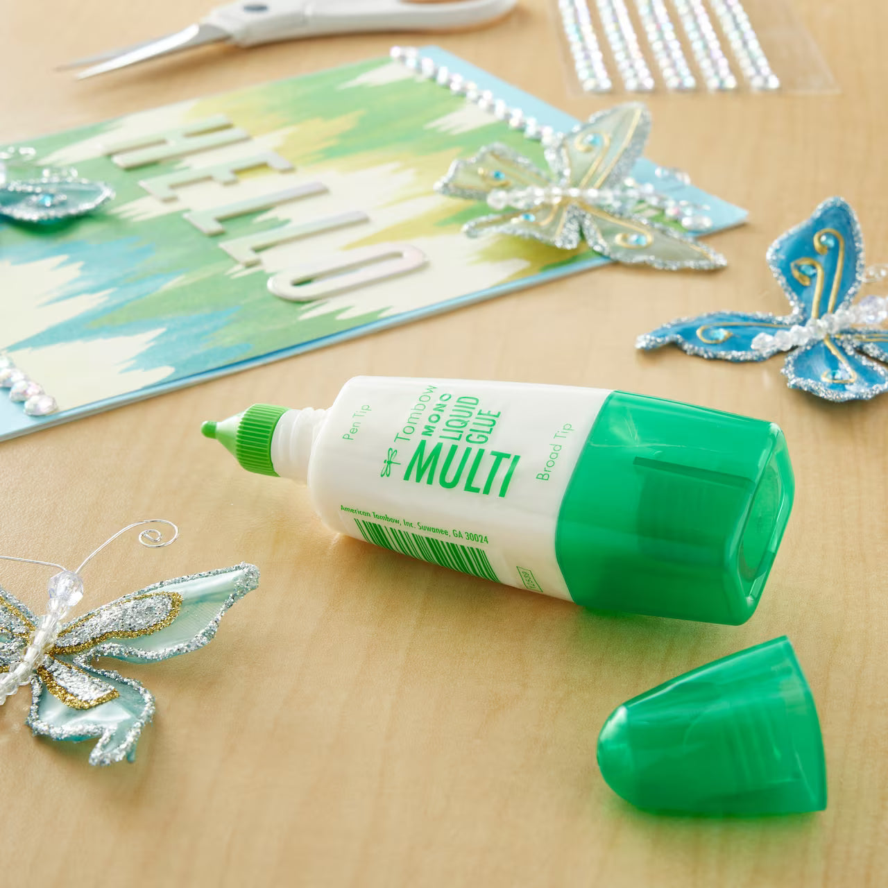 Bottle of Tombow Multi Liquid Glue on a wooden surface with decorative butterflies and cards.