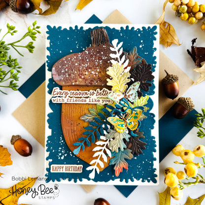A handmade layered greeting card featuring an acorn, autumn leaves, a butterfly, and decorative text, styled on a white surface with scattered acorns, leaves, and paper.