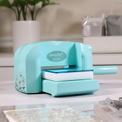 A turquoise paper cutter machine with floral accents sits on a light countertop in a kitchen or craft workspace, with blurred cabinets, a plant, and kitchen items in the background.