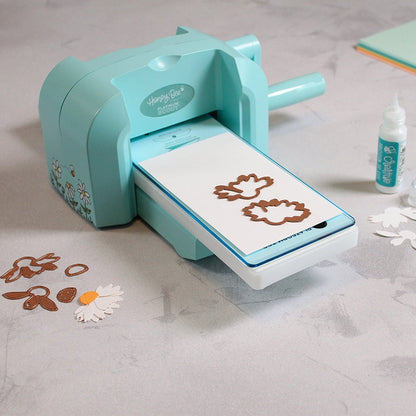 Bee Creative Platinum Scout die cutting machine with die-cut pieces on the plates, flower pieces and glue on the table