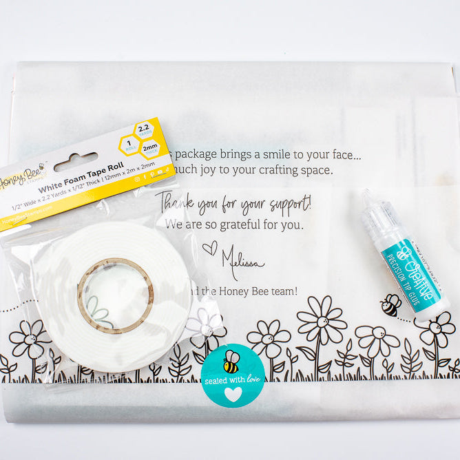 A white floral honey bee craft kit on a white background, including a clear envelope, a roll of white washi tape, a small tube of white glue, and a Honey Bee package.