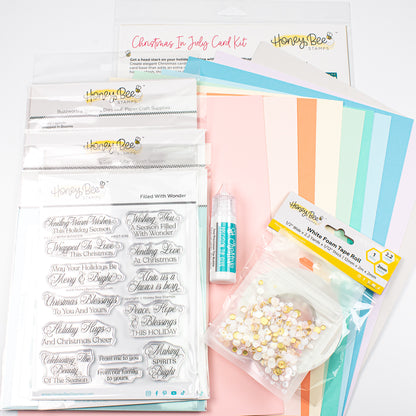 A collection of craft supplies from Honey Bee Papercraft, including a Christmas in July card kit, white flower tape buds, clear stamps, and pastel striped cardstock sheets, arranged on a white background.