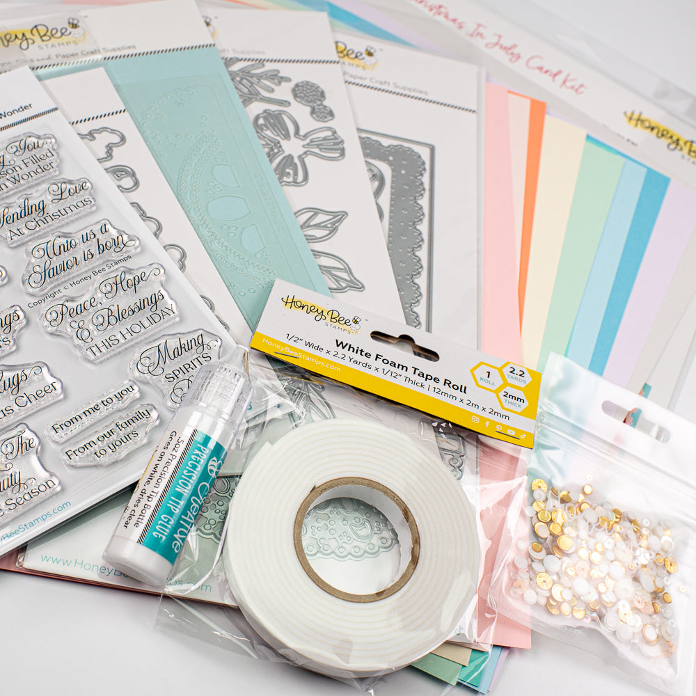 A craft kit assortment laid out on a white surface, including a roll of white foam tape, clear stamps, metal dies, pastel cardstock sheets, a tube of adhesive, and a bag of decorative pearls. The packaging shows the brand Honey Bee.
