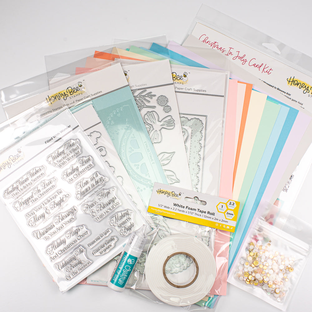 A craft kit bundle from Honey Bee Supplies featuring a Christmas in July card kit, clear stamp sheets with holiday phrases, a white foam tape roll, metal dies, and a small bag of pearl embellishments on a light gray background.