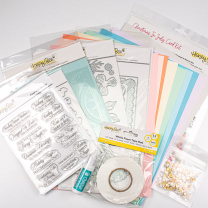 A craft kit bundle from Honey Bee Supplies featuring a Christmas in July card kit, clear stamp sheets with holiday phrases, a white foam tape roll, metal dies, and a small bag of pearl embellishments on a light gray background.