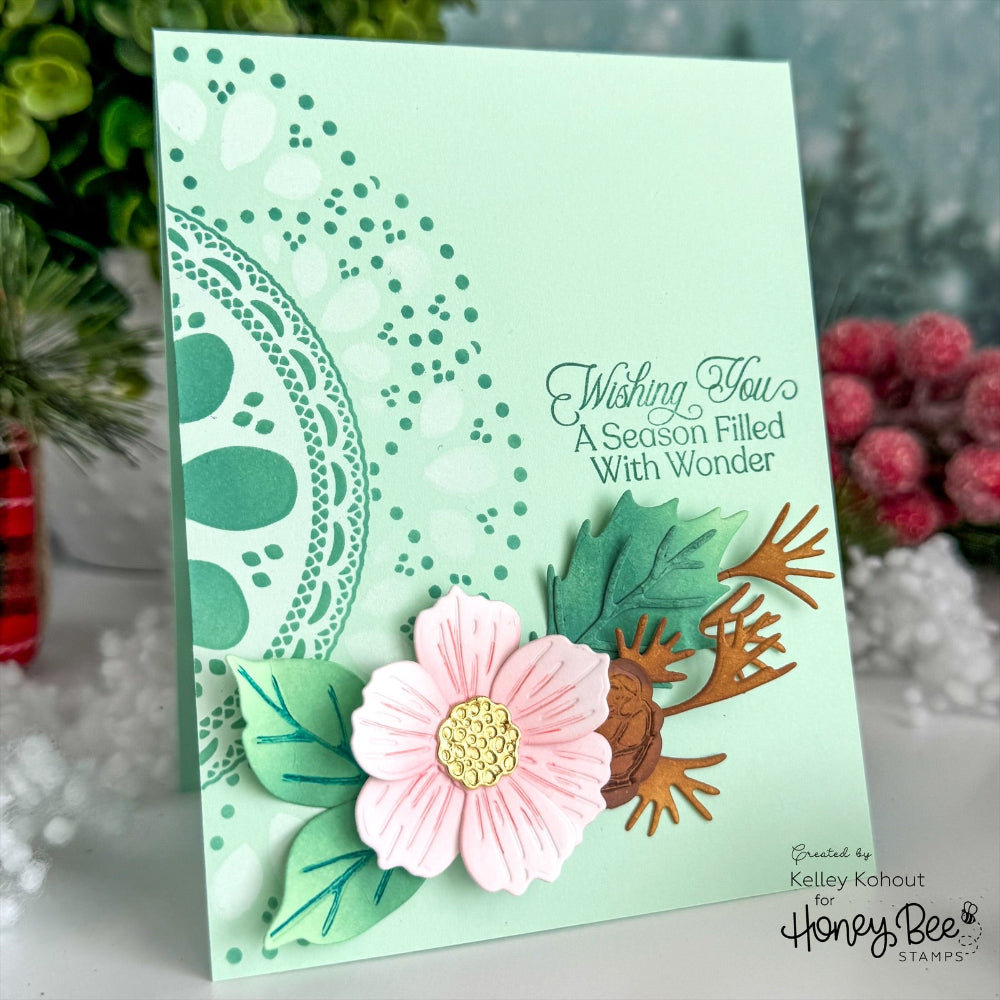 A handmade greeting card with a mint green background, featuring a pink paper flower, green layered leaves, and brown paper birds. The card has stamped text reading 'Wishing You A Season Filled With Wonder' and is photographed standing upright on a white surface with blurred greenery and pink flowers in the background.