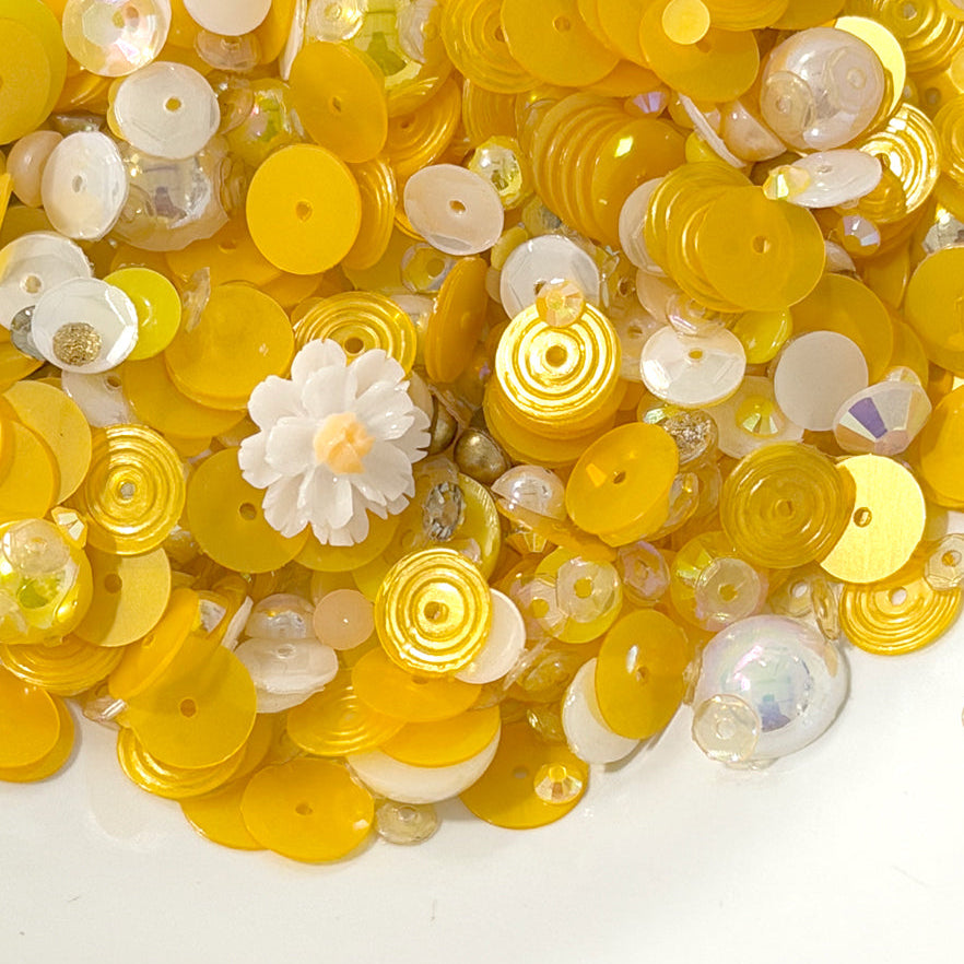 A pile of yellow and white craft sequins, beads, and small decorative flower pieces on a plain white background.