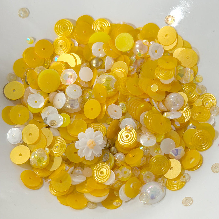 A pile of yellow and white craft sequins, beads, and small decorative pieces on a plain white background.