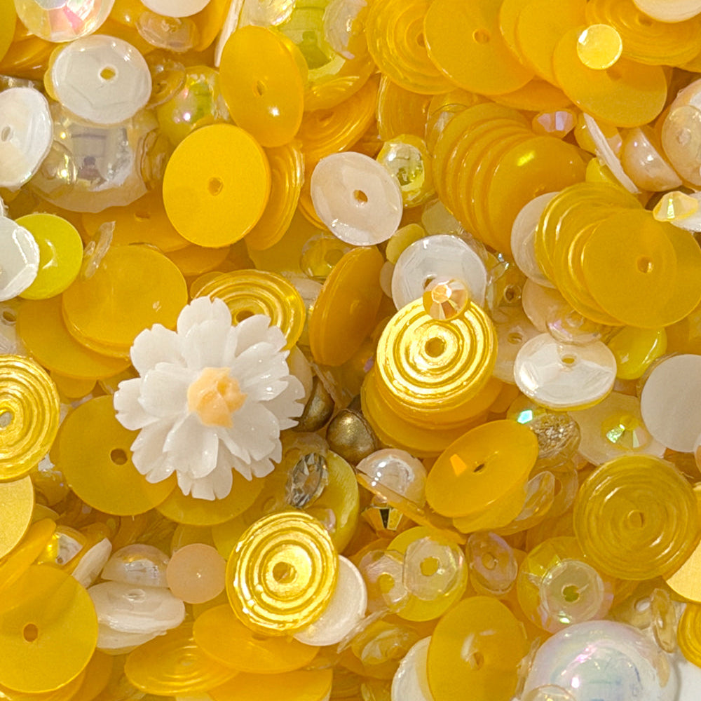 A close-up pile of craft embellishments, including many translucent yellow and white round sequins, spiral disc beads, clear faceted beads, and a small white flower-shaped bead.