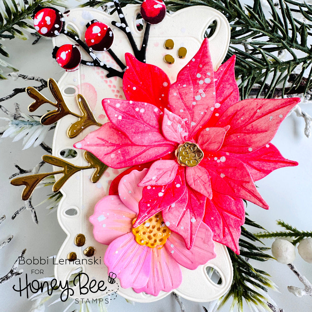 A decorative floral embellishment featuring two poinsettia-style flowers in bright pink and coral-red tones with white speckles, gold centers, and a white backing, styled with faux pine branches and red berries. Text in the lower left reads 'Bobbi Lemanski for Honey Bee STAMPS'.