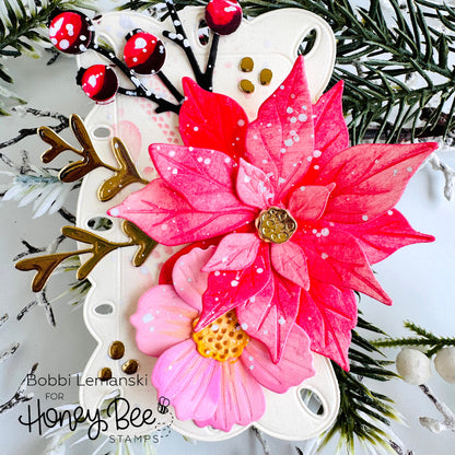 A decorative floral embellishment featuring two poinsettia-style flowers in bright pink and coral-red tones with white speckles, gold centers, and a white backing, styled with faux pine branches and red berries. Text in the lower left reads 'Bobbi Lemanski for Honey Bee STAMPS'.