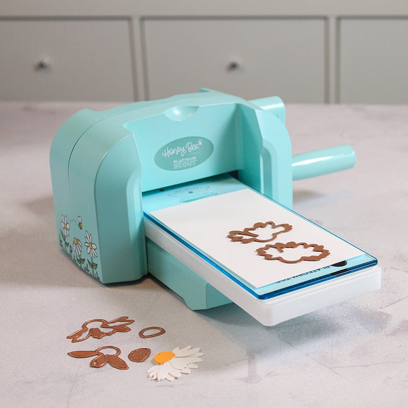 A light blue die-cuting machine on a light gray marble countertop, with die-cut paper shapes emerging from the slot. A small bottle of adhesive and loose paper pieces are placed nearby, with a crafting mat visible in the background.