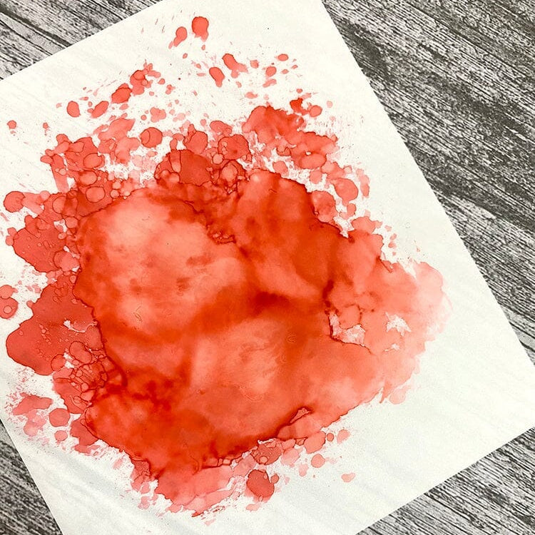 Red watercolor stain on a white paper sheet with a gray wooden background