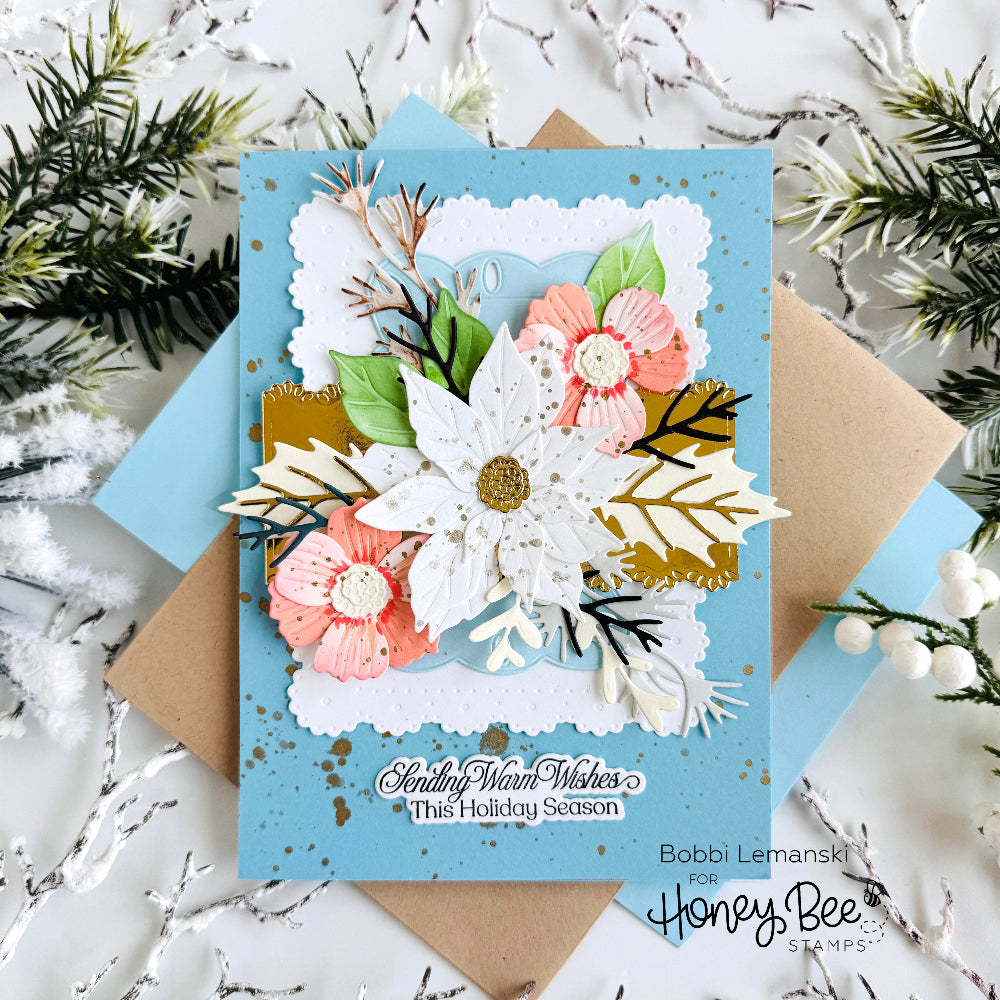 A handmade layered holiday greeting card with a light blue base, featuring white and pink paper flowers, green leaves, and gold foil accents. The card is layered over kraft paper and blue envelopes, styled with white pine branches and silver tinsel on a white marble background.