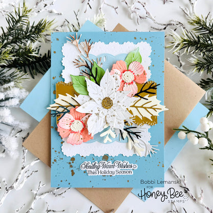 A handmade layered holiday greeting card with a light blue base, featuring white and pink paper flowers, green leaves, and gold foil accents. The card is layered over kraft paper and blue envelopes, styled with white pine branches and silver tinsel on a white marble background.