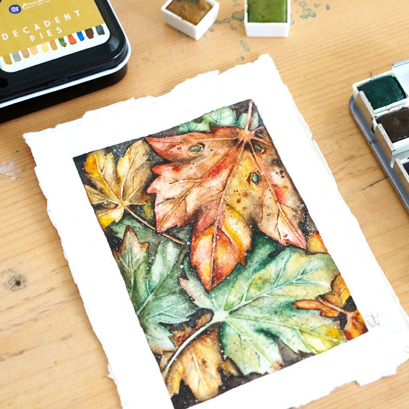 Watercolor painting of leaves on paper with watercolor set and ink pad on wooden surface