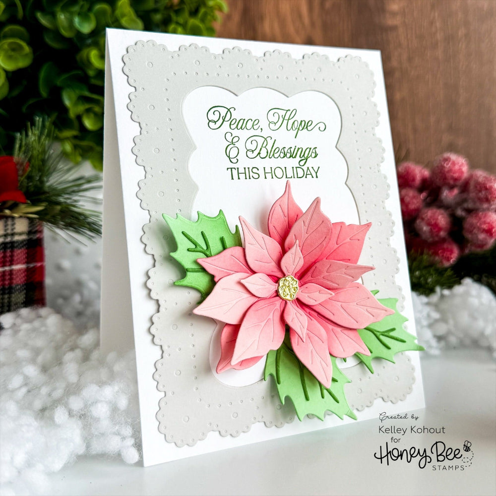 A handmade holiday greeting card featuring a layered pink poinsettia with green leaves and a scalloped white border. The card is displayed upright in a festive setting with blurred greenery, white faux snow, and red berries in the background.