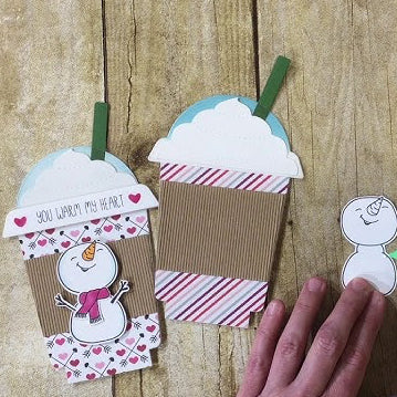 Hand applying glue to a snowman cutout next to paper coffee cups on a wooden surface.