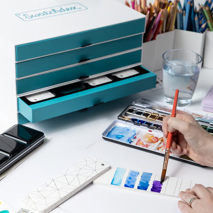 Person using a watercolor set with a blue watercolor box and a glass of water on a white surface.